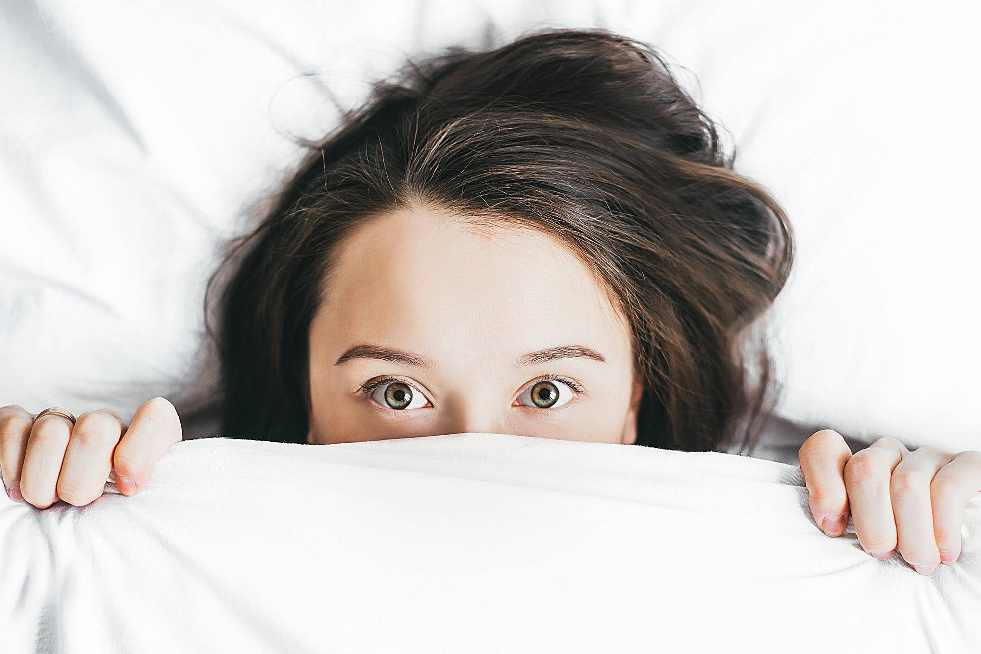 A young brunette woman laying in bed face-up with the covers drawn up over her nose, looking frightened