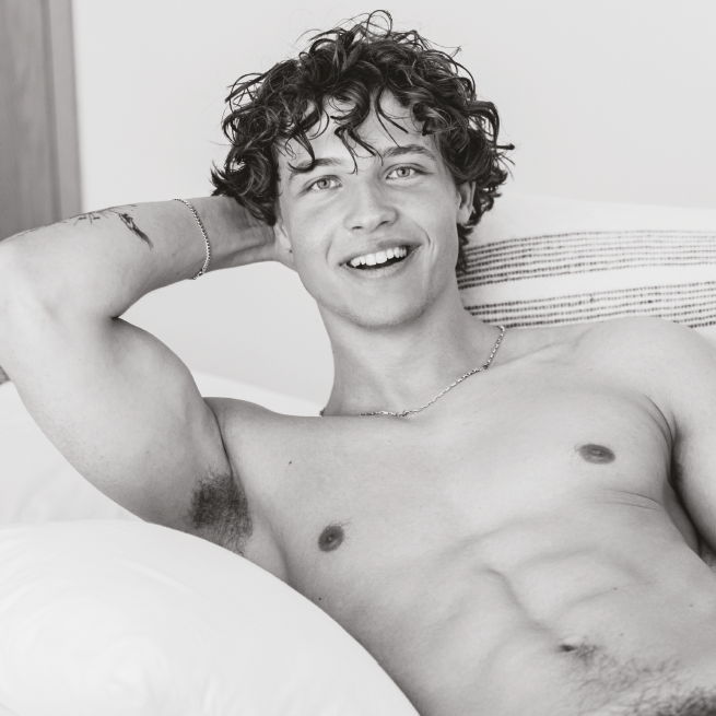 Young man with curly hair smiling, sitting shirtless on a couch.