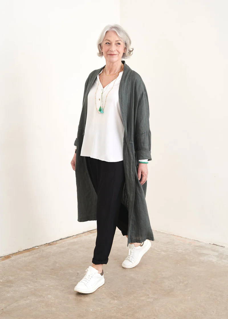 A model wearing a long blue grey linen trnech coat over a white top, black trousers and white trainers