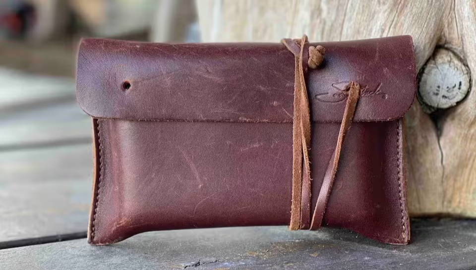 Dark brown full-grain leather wallet with natural scuffs and a tied leather cord leaning against rustic weathered wood.