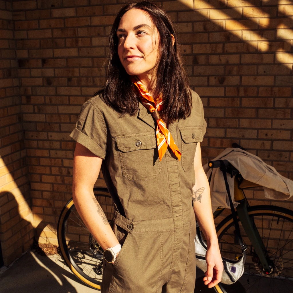 Person in an olive jumpsuit with a scarf, standing against a brick wall.
