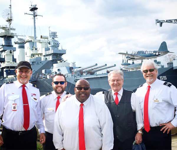 A group of bus drivers in uniform with red ties posing in front of a warship outside