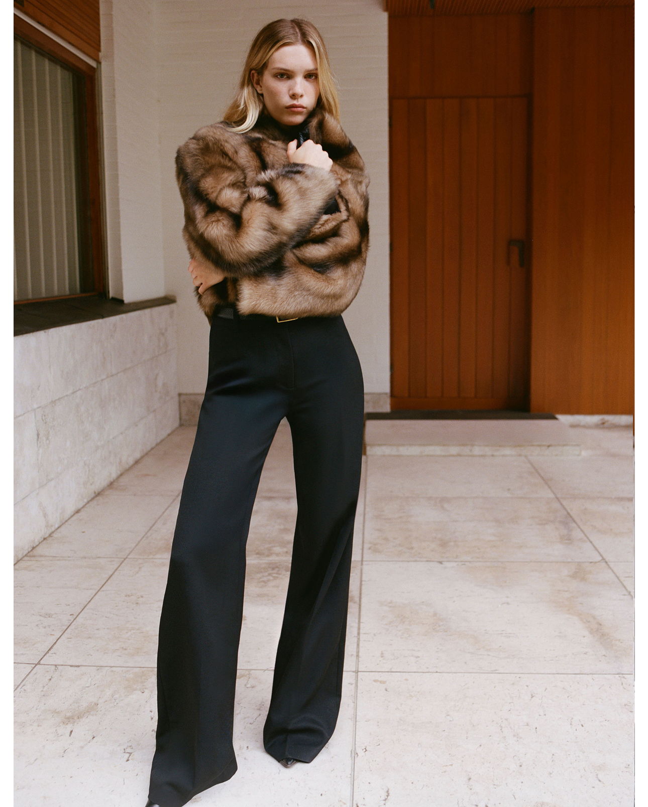 Model in a stylish fur coat and black trousers posing indoors.