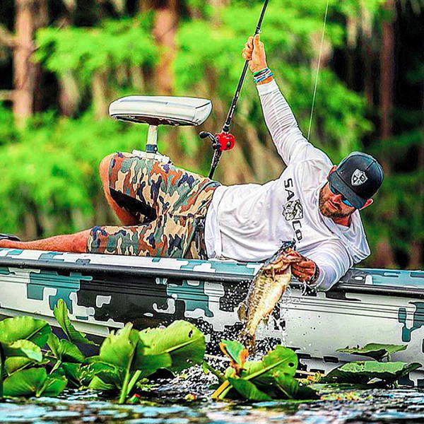 Ryan Copenhaver laying on a boat pulling a fish out of the water wearing a hooded performance shirt and an SA Company branded hat.