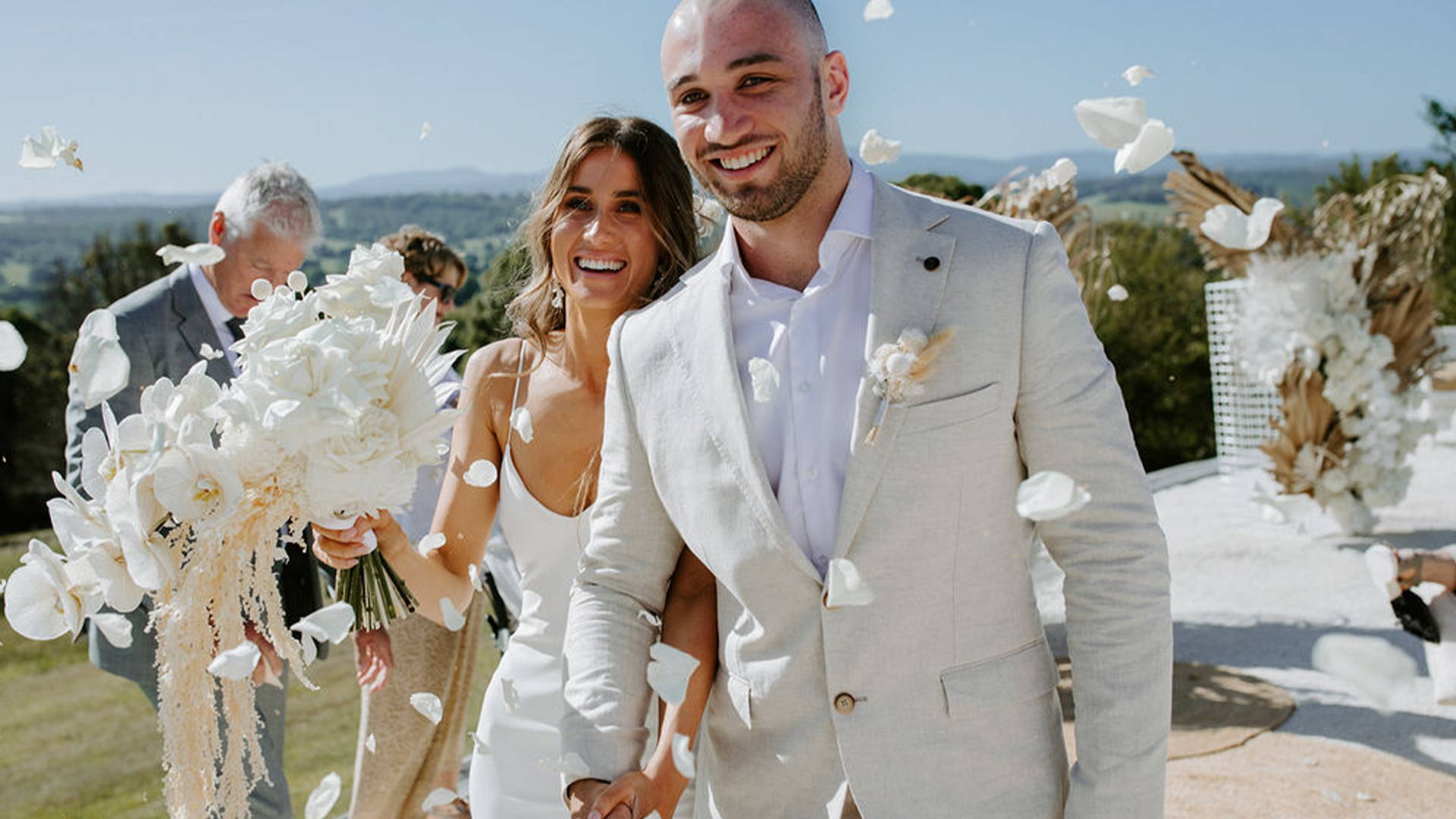 Groom wearing a beige suit walking with his bride with white flowers