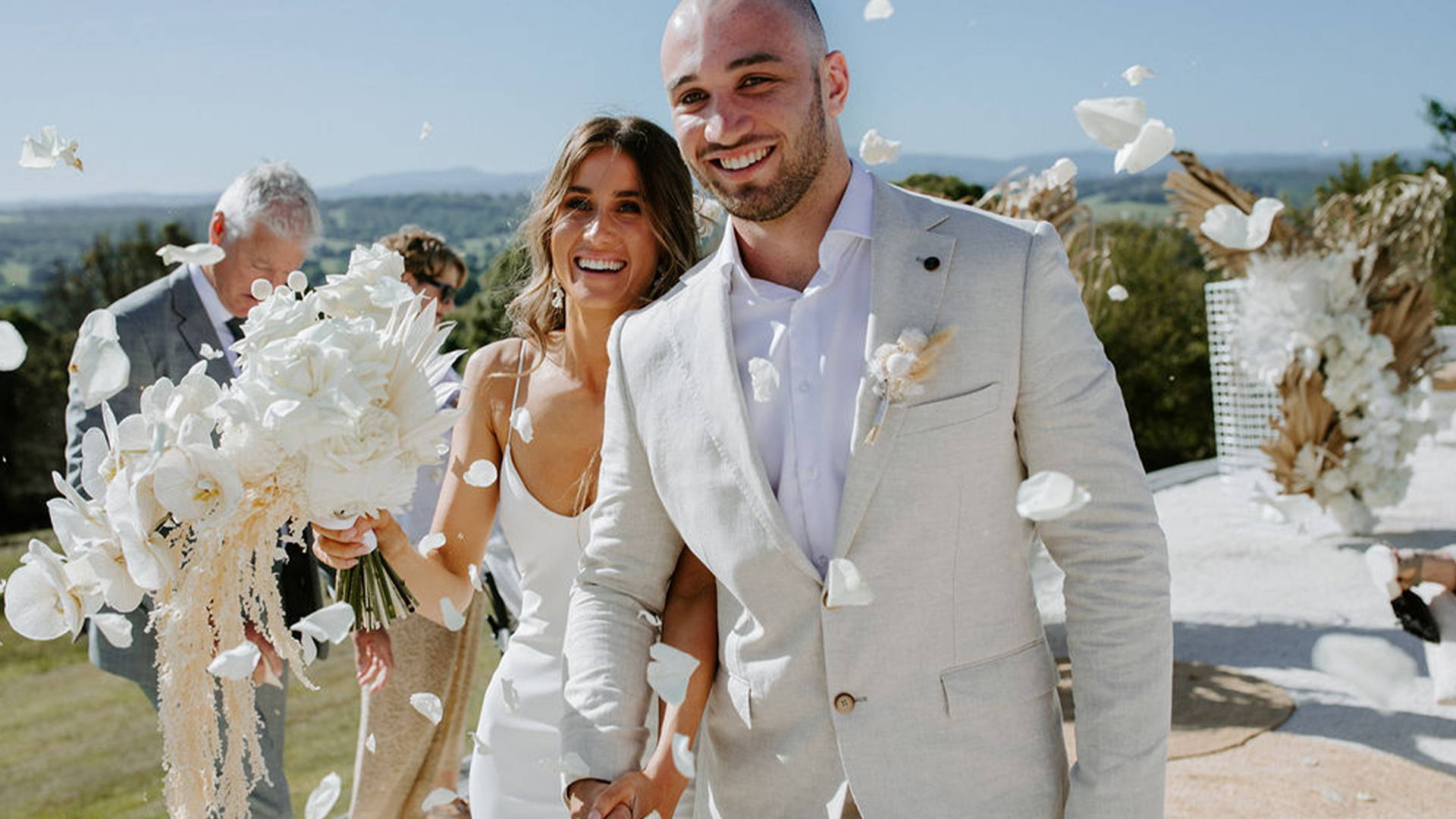 Novio con traje beige caminando con su novia con flores blancas