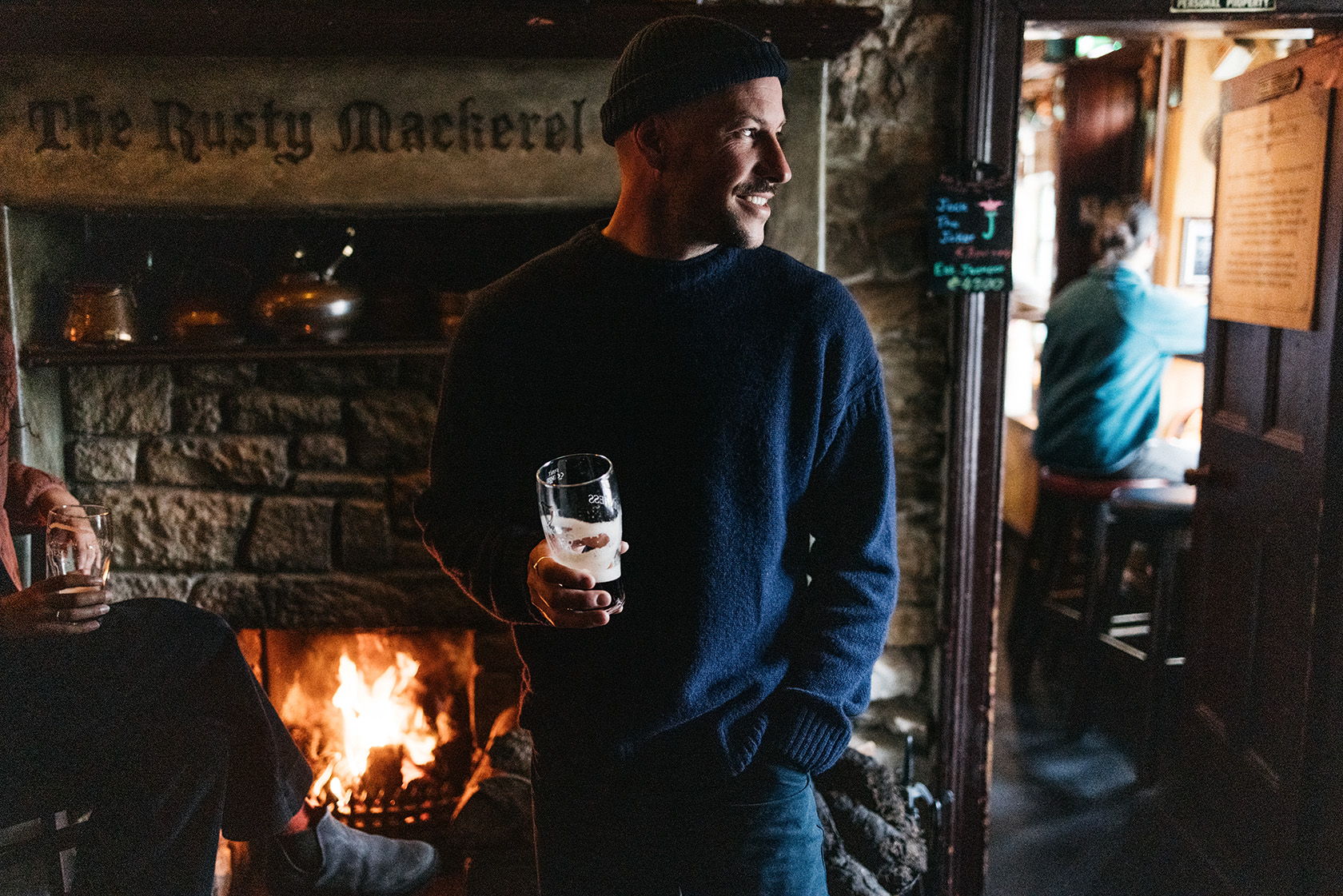 Man holding a drink, standing by a cozy fireplace in a pub.