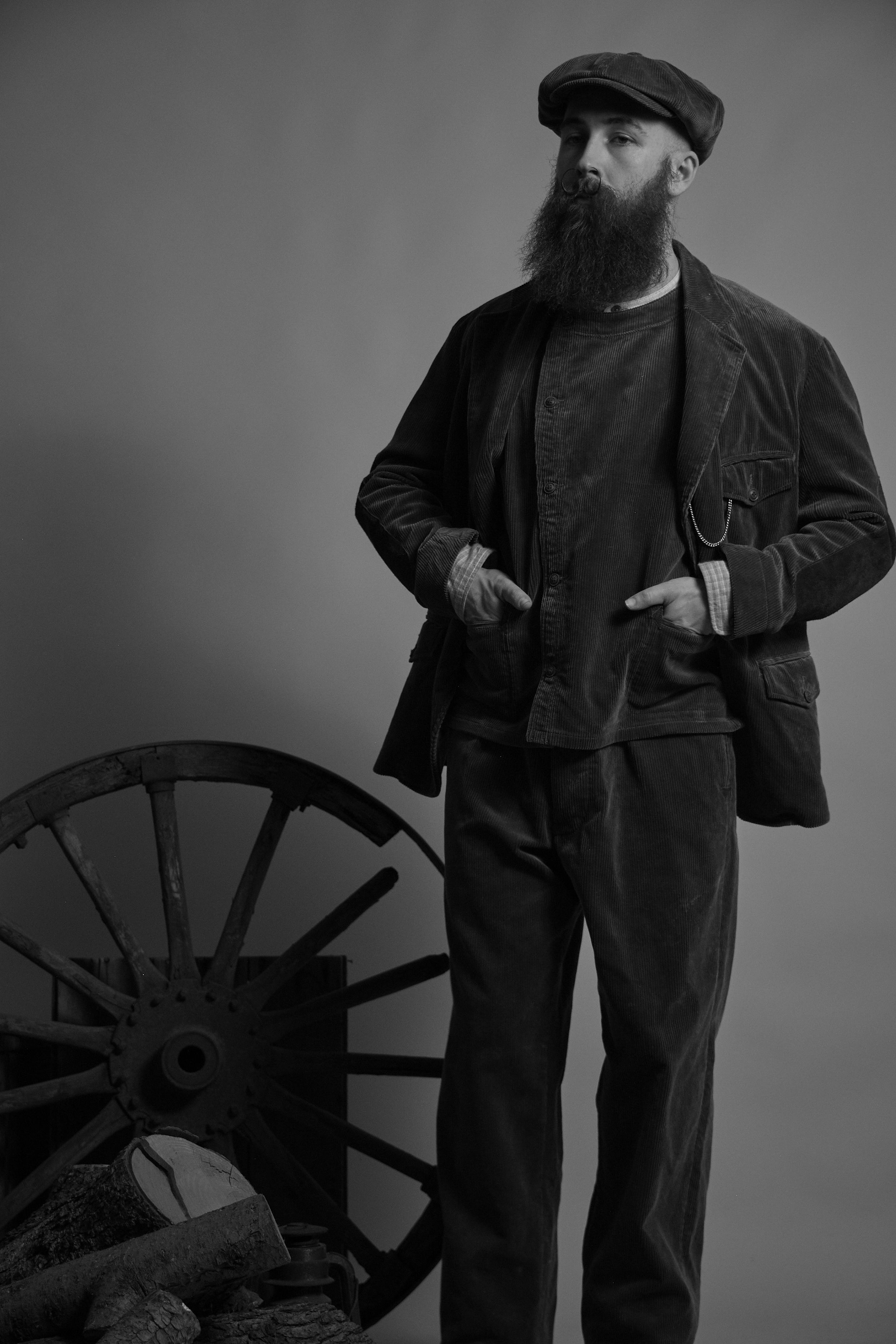 A man with a beard wearing a cap and vintage clothing stands beside a wheel.