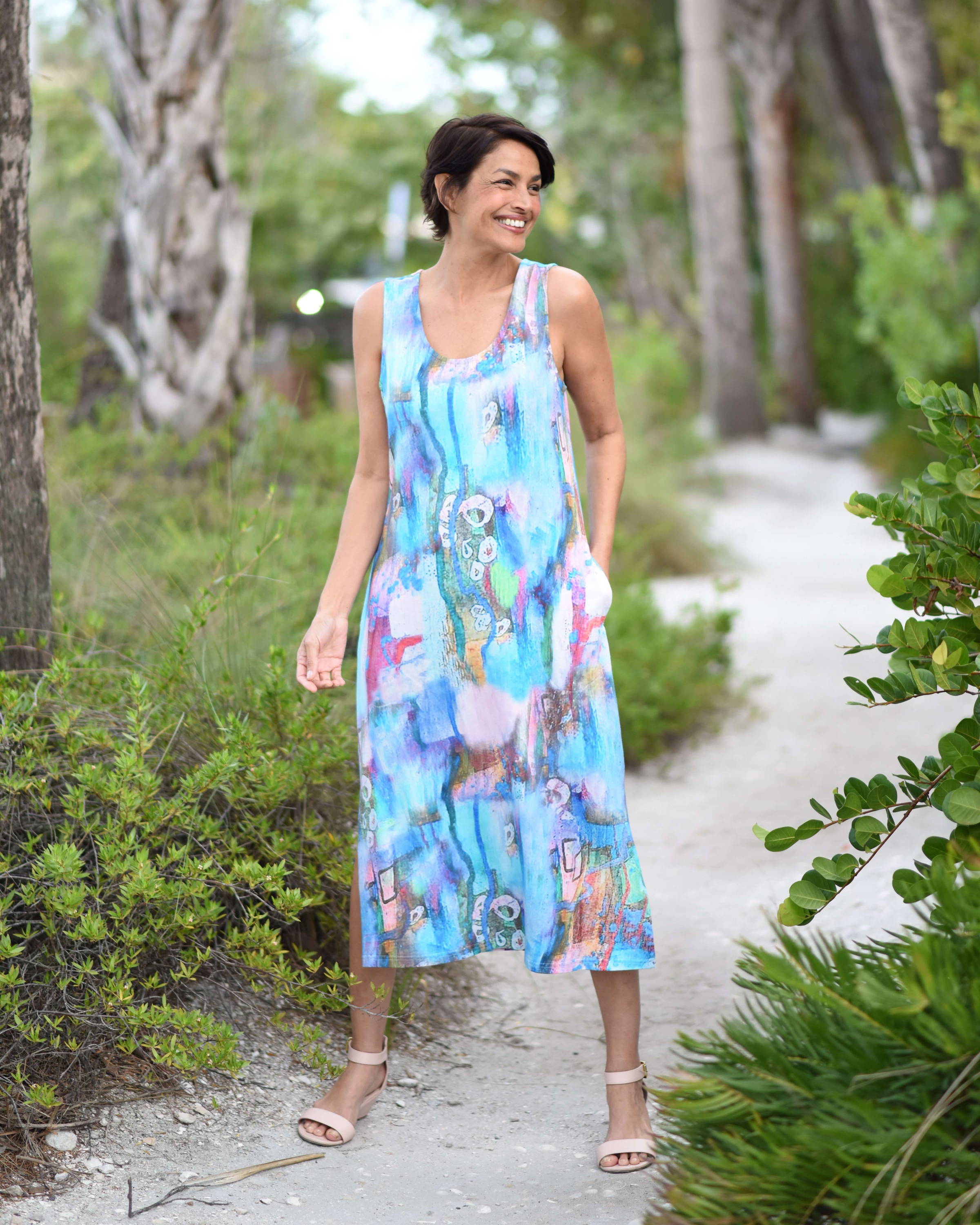 A smiling woman with short dark hair wearing a sleeveless pastel-colored dress walks along a nature path. . 