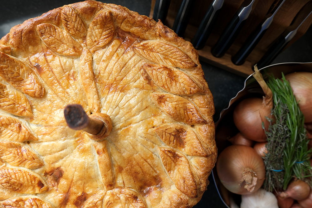 A golden-brown lamb shank pie beside a bowl of onions and herbs.