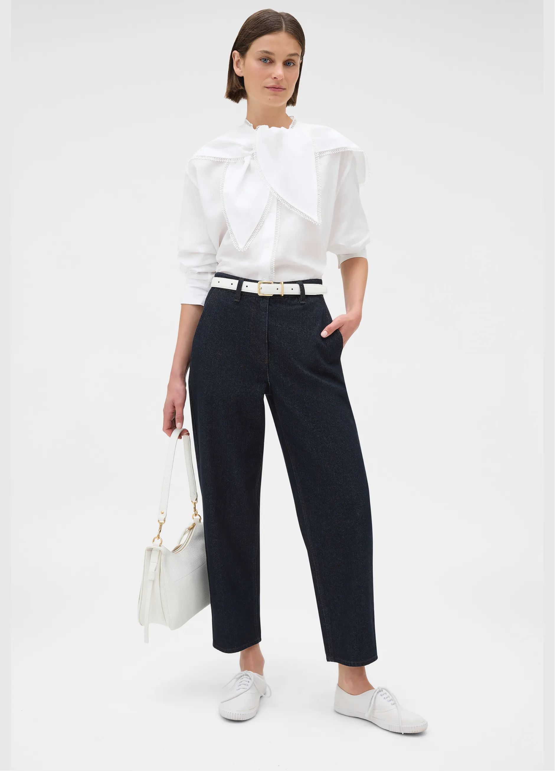 Model wearing a white blouse, navy pants, and holding a white handbag.