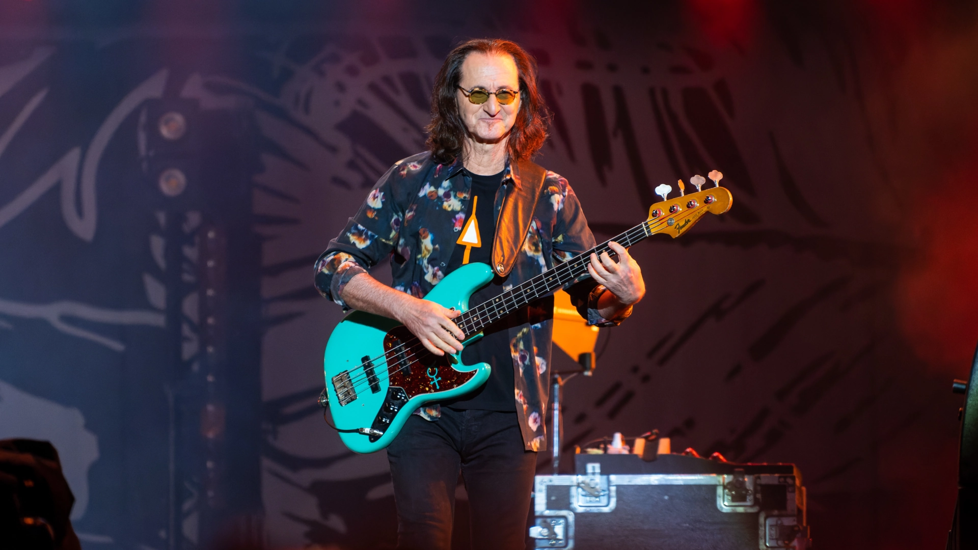 Geddy Lee of Rush playing a turquoise Fender Jazz Bass on stage with dramatic lighting.