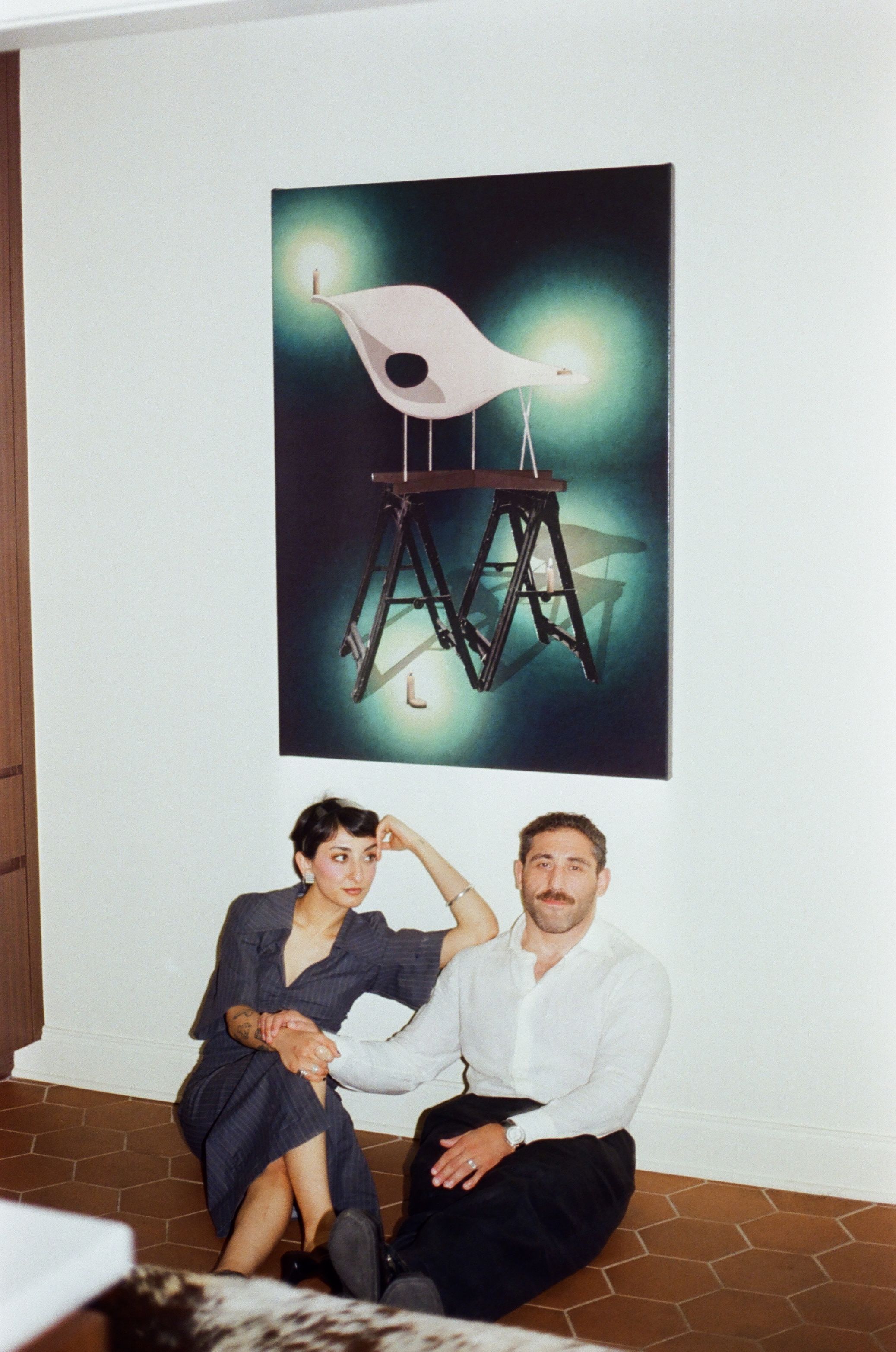 A couple sitting on the floor beside a chair artwork on the wall.