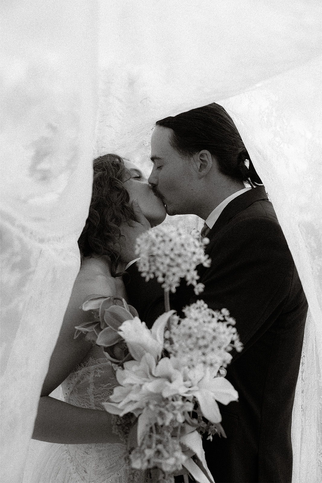 Couple kissing under a veil, holding wedding flowers. Black and white photo.