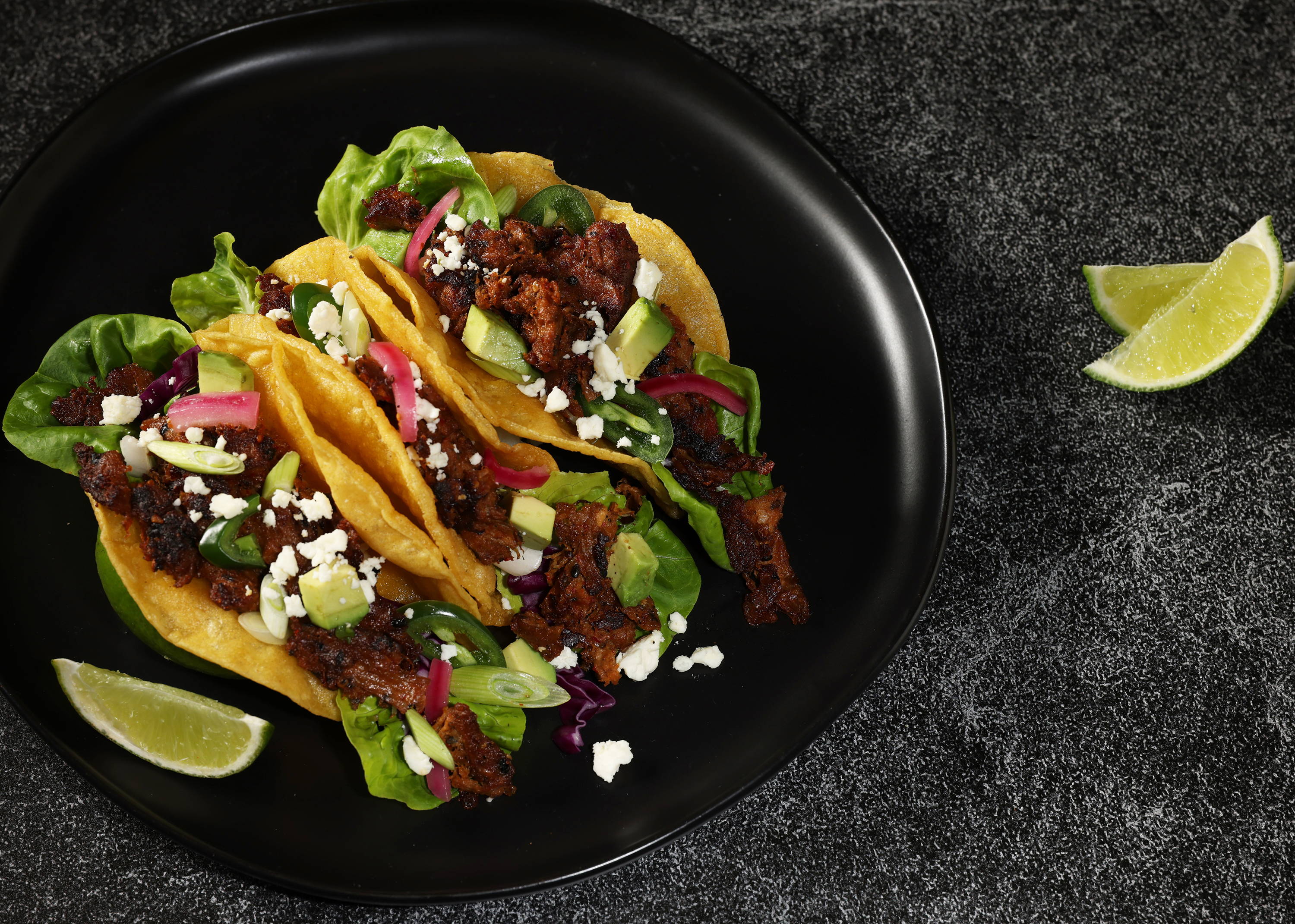 Mexican Barbacoa Harvest Shreds served in three corn taco shells with lettuce, pickled red onions, avocado chunks and queso fresco. On a black plate with lime garnish