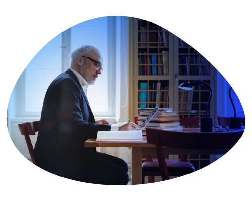 Elderly man reading at a table in a library with bookshelves.