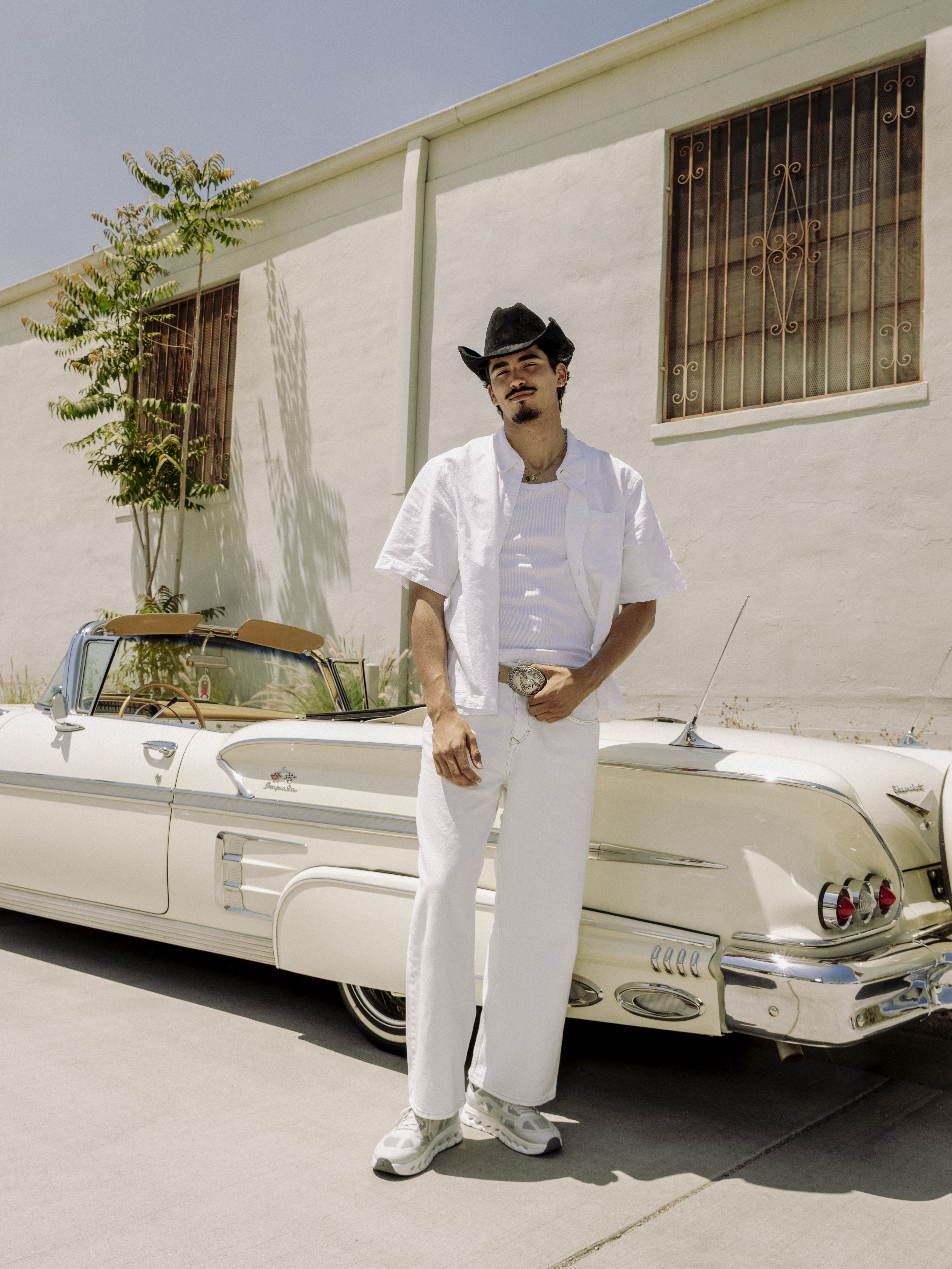 Person in white outfit standing beside a vintage car in a sunny location.