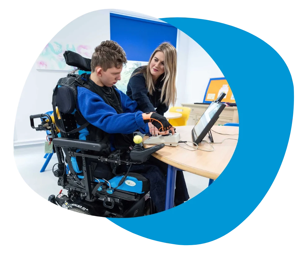 A young boy in a wheelchair receives assistance at a table.