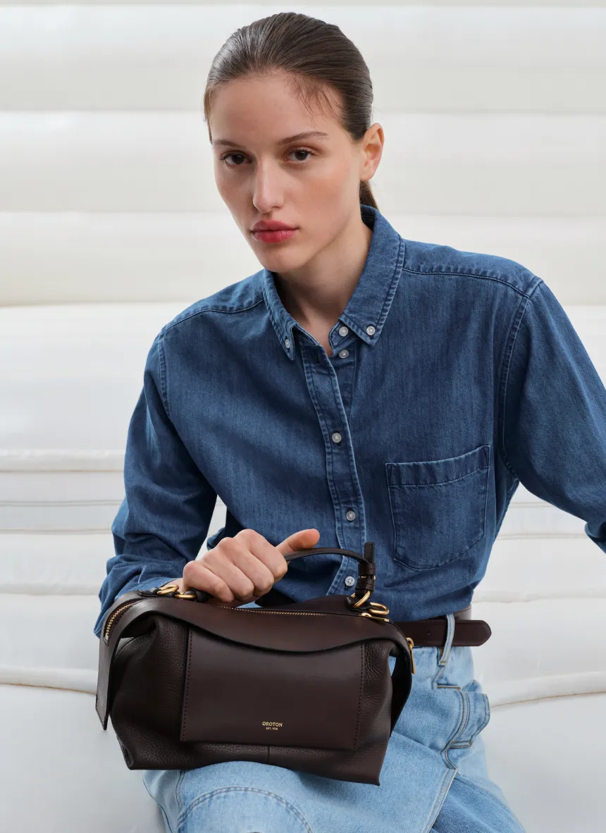 Woman in denim shirt holding a brown handbag.