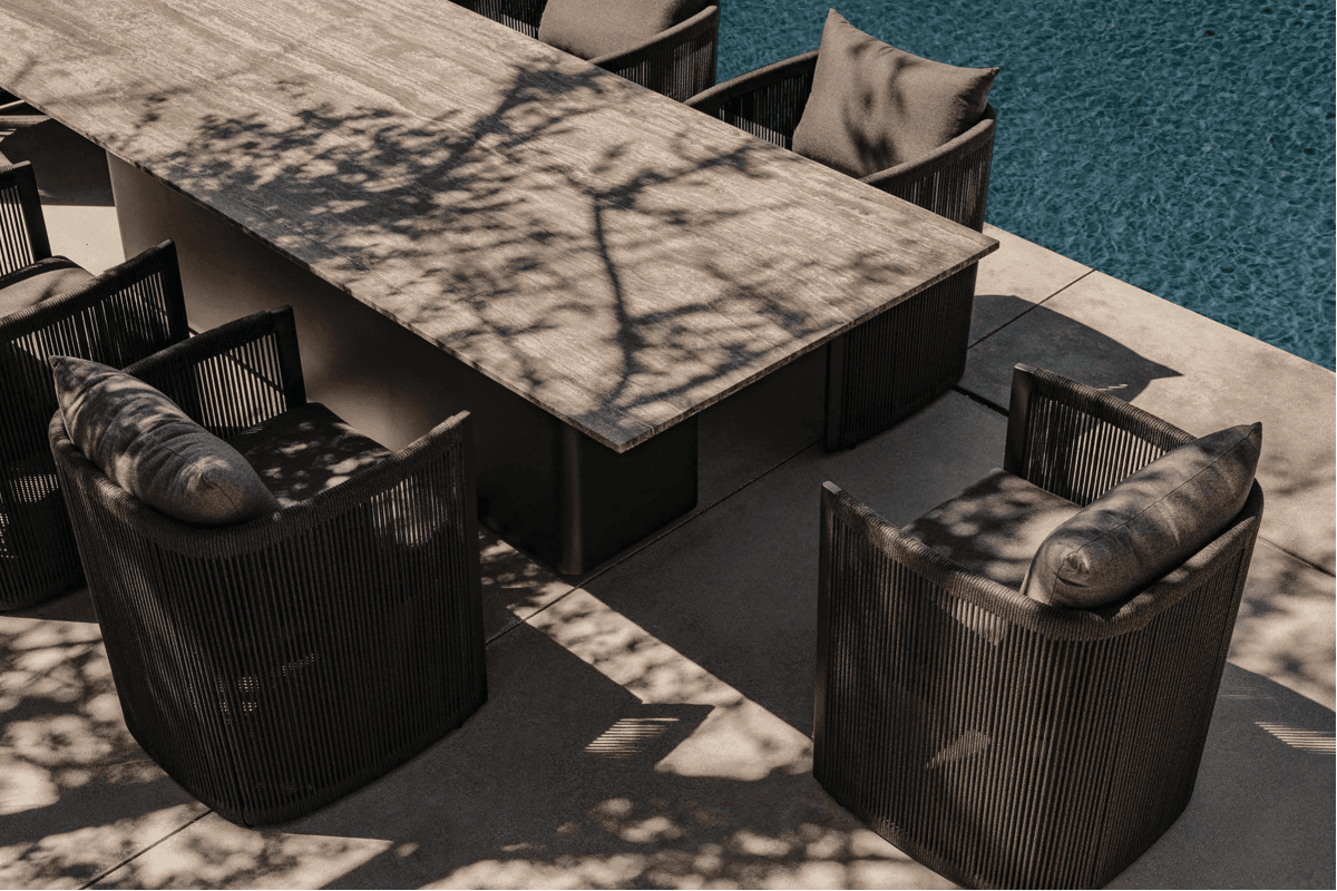 Close-up of a dark outdoor dining table and black rope-wrapped dining chairs beside a swimming pool, with tree shadows cast across the surface.