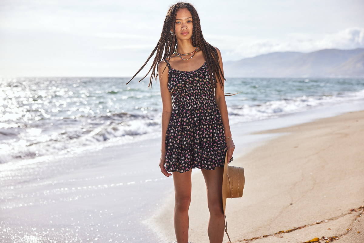 Trixxi sun kissed summer lookbook link, girl on beach holding a sun hat in a floral tank dress.