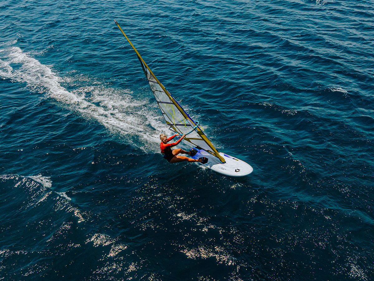 A person windsurfing on blue water, creating waves behind them.