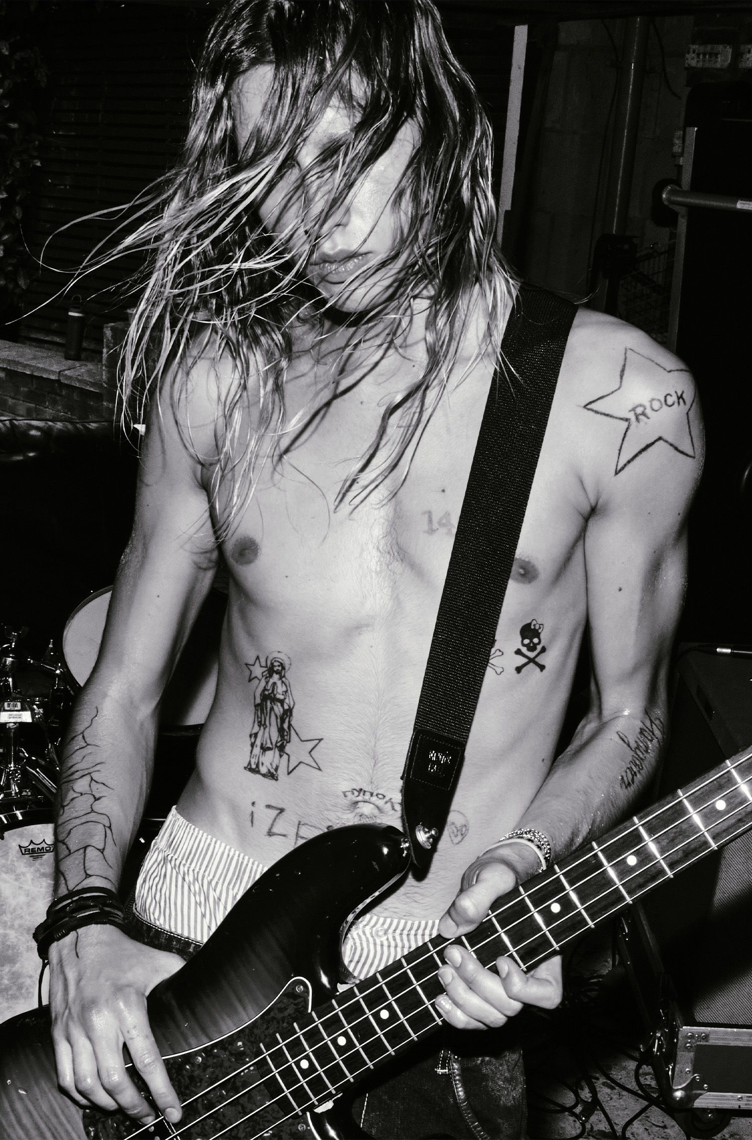 Shirtless musician with long hair playing a bass guitar on stage.