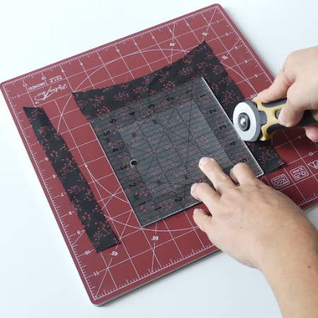 Cutting a dark fabric piece on a rotating cutting mat with cutter and square ruler