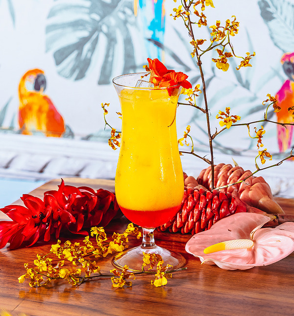 A colorful cocktail garnished with flowers and tropical fruits on a wooden table.
