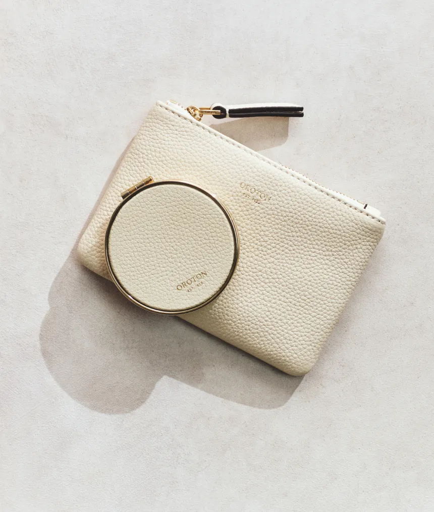 A cream-colored pouch with a circular mirror beside it on a light background.
