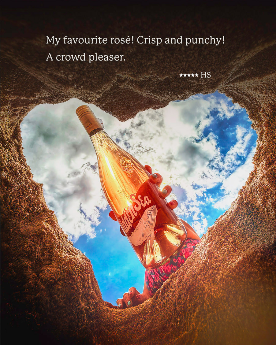 A hand holding a The Hidden Sea rosé bottle up through a heart-shaped opening in sand with a customer quote: “My favourite rosé! Crisp and punchy! A crowd pleaser.” — 5-star review.