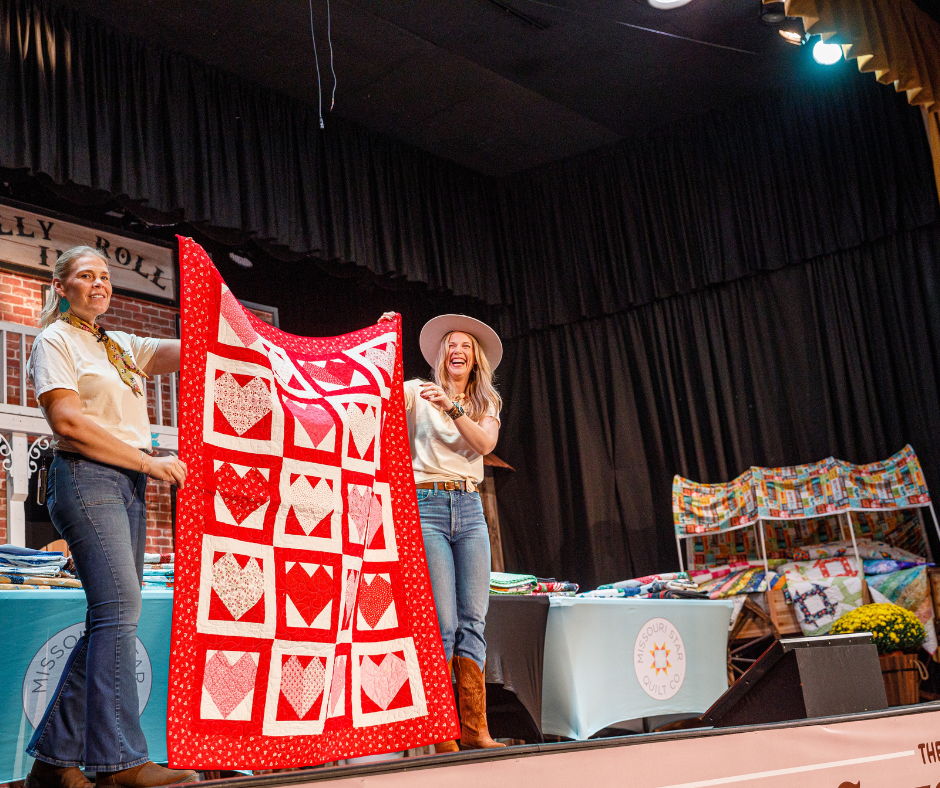 Two women display a red quilt on stage at an event.