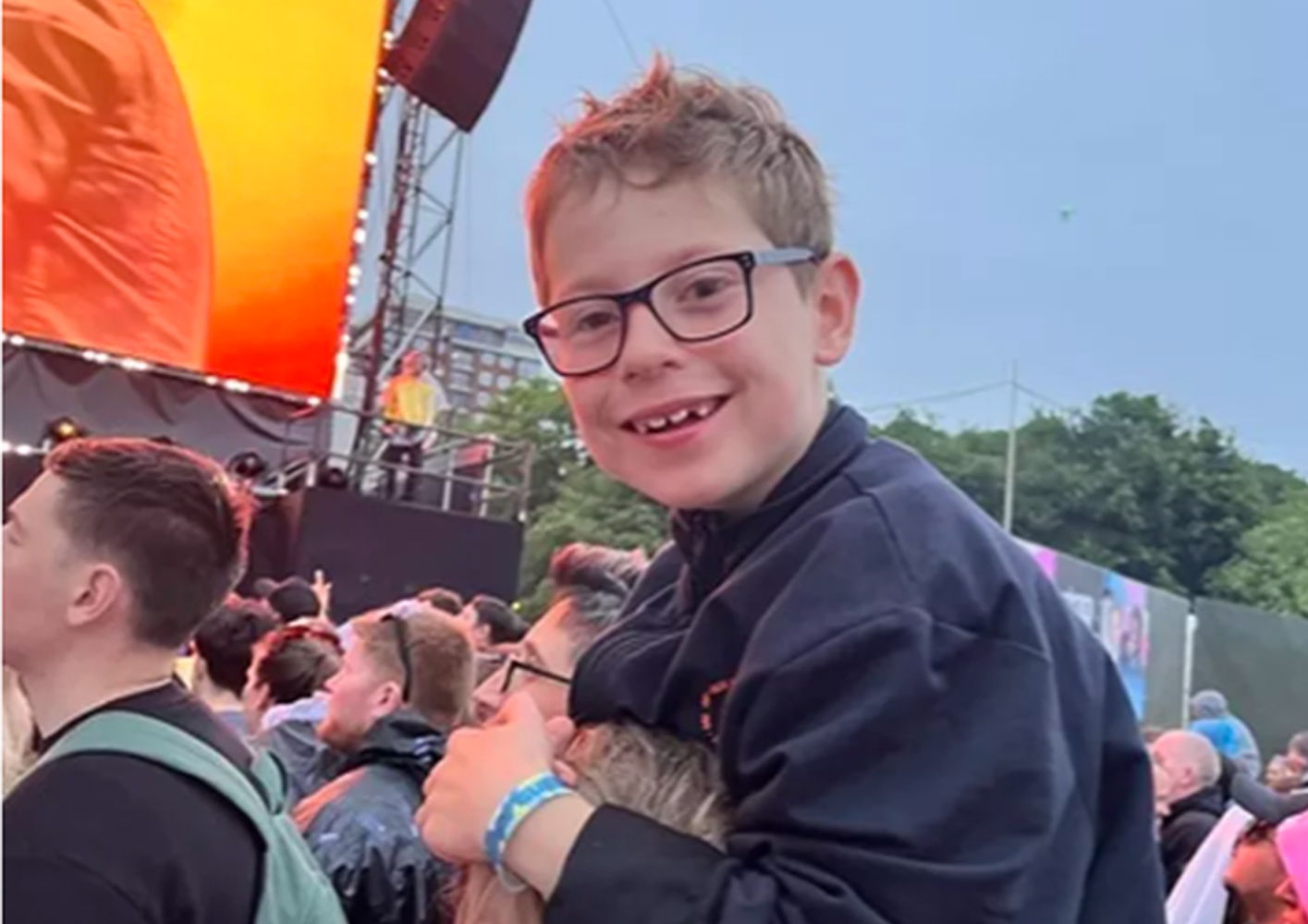 Boy with glasses smiling on someone's shoulders at a concert.