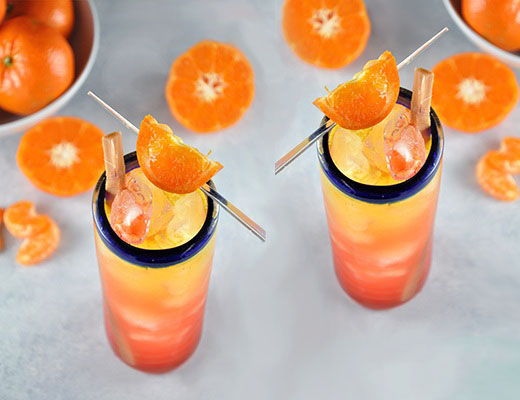Two colorful cocktails garnished with orange slices, surrounded by tangerines.