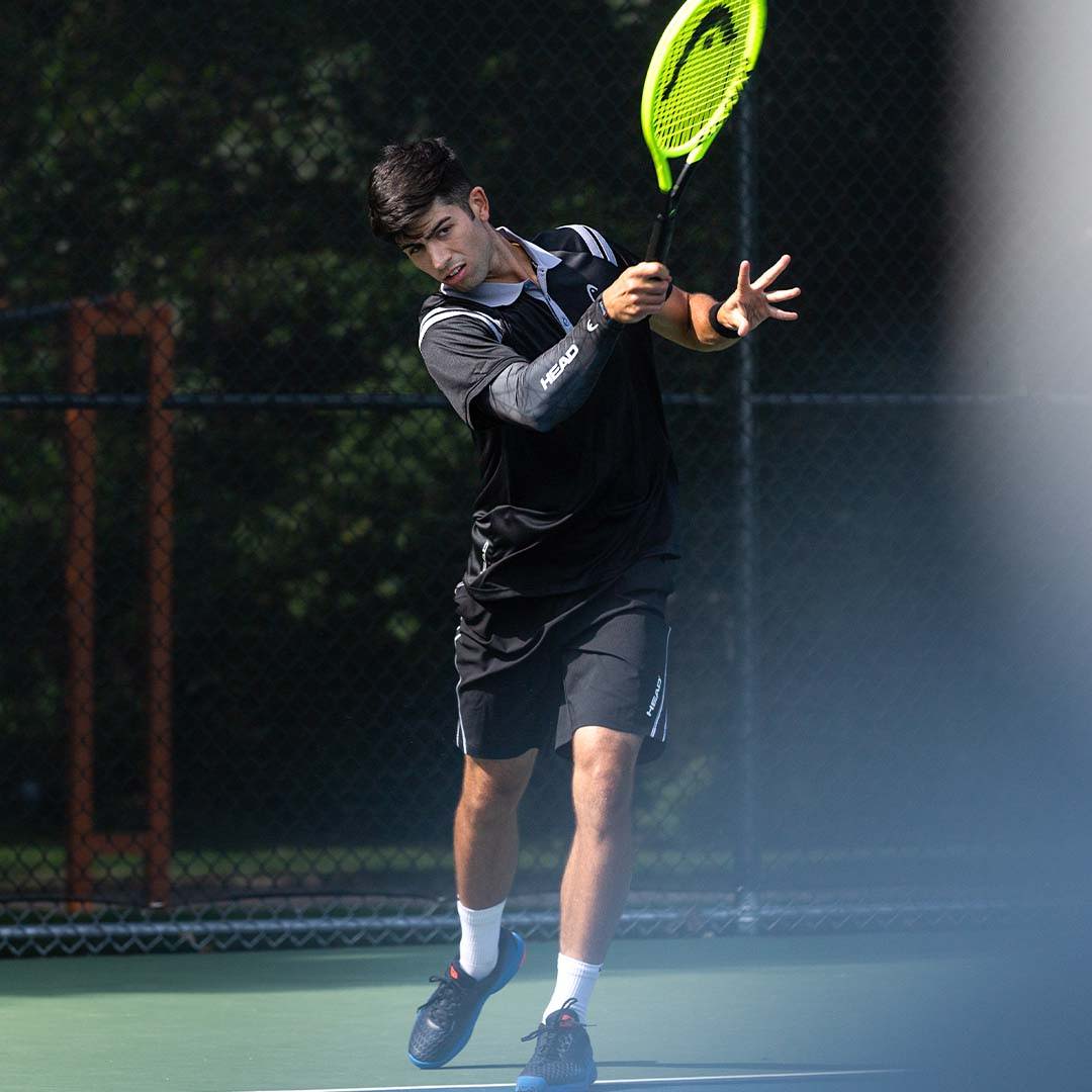 A tennis player swings his HEAD™ tennis racquet confidently, wearing an eSmartr x HEAD™ smart compression sleeve on his swinging arm.