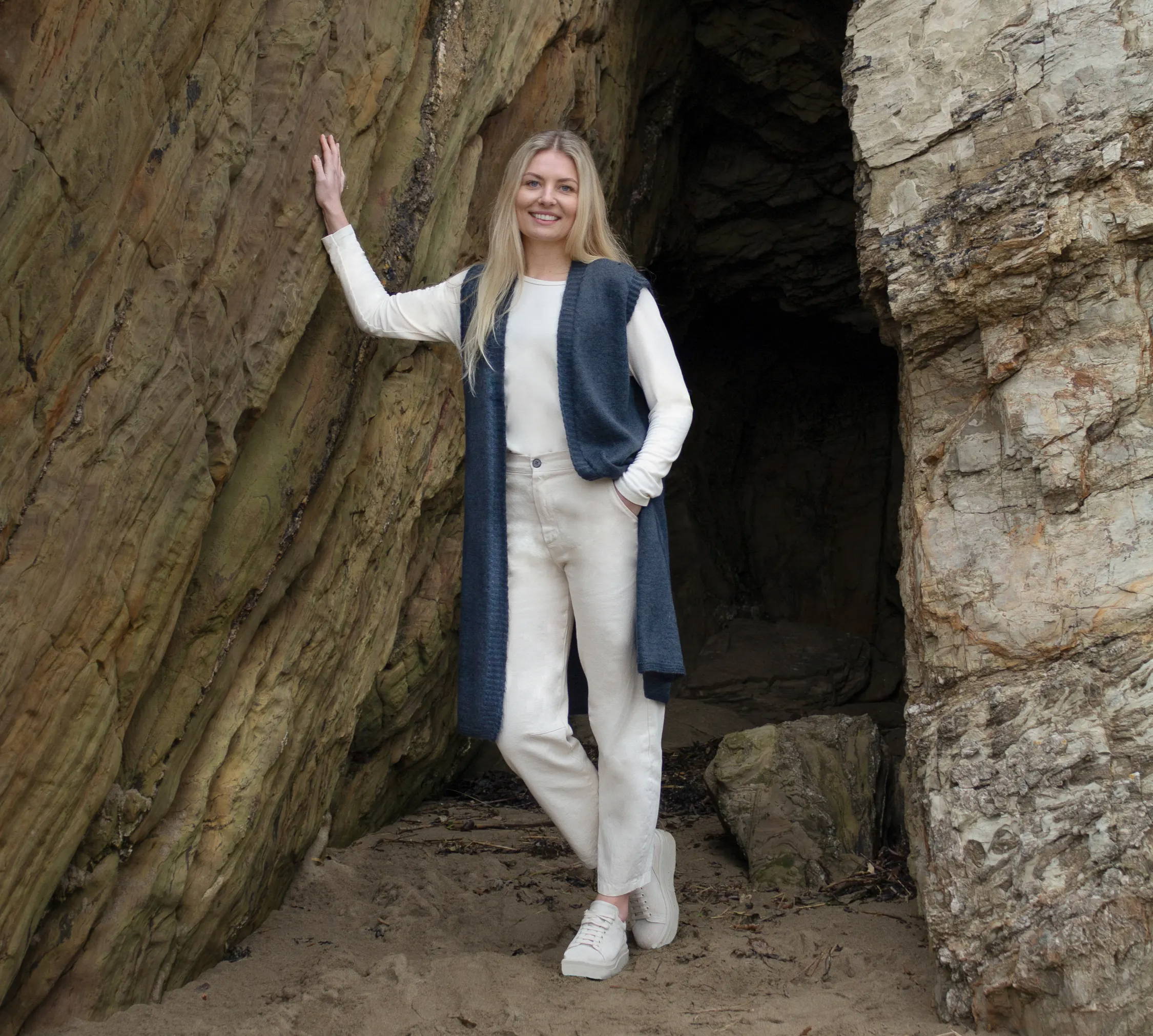 A model on a beach wearing a white long sleeved top, beige trousers, beige trainers and a long blue knitted sleeveless cardigan