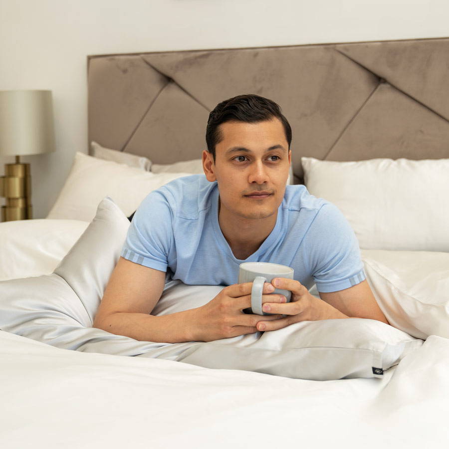 man holding cup of coffee resting on grey silk pillowcase