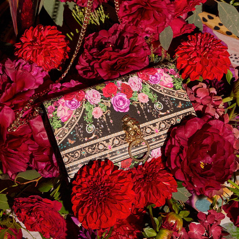 CAMILLA RED BLACK FLORAL CLUTCH BAG LAYING IN BED OF RED FLOWERS