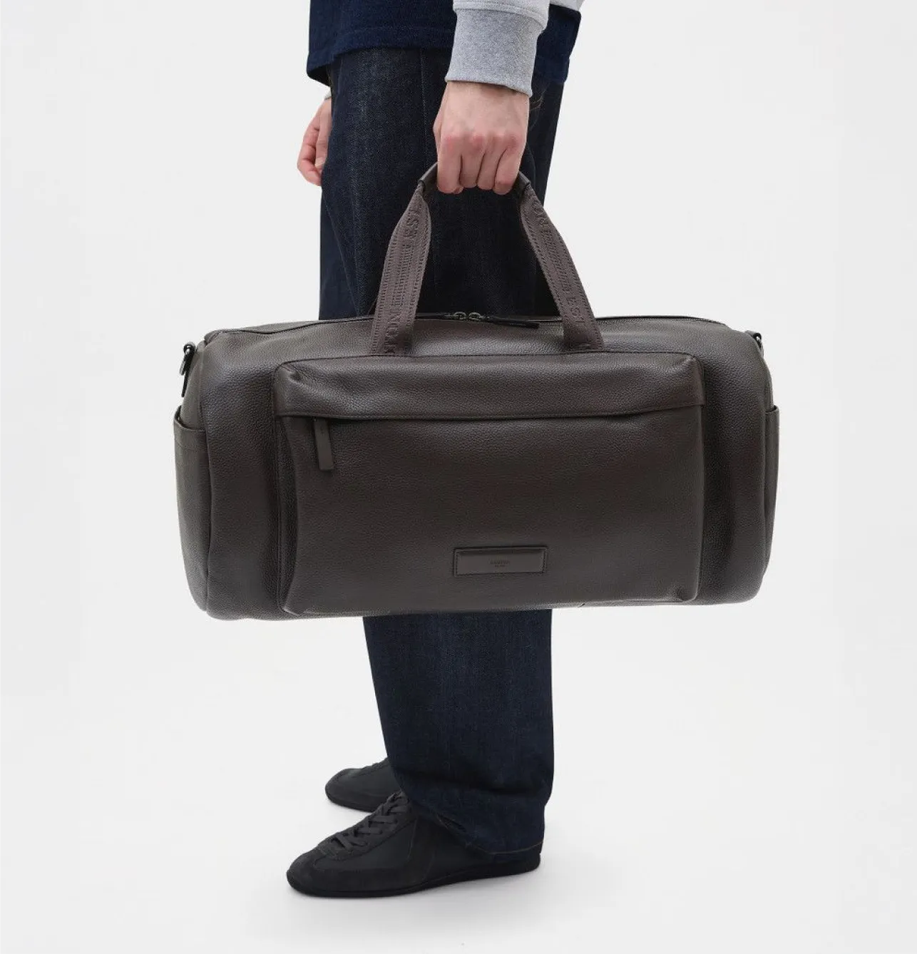 Person holding a dark brown duffle bag against a white background.