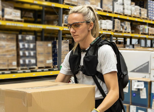 Person in a warehouse lifting a box while wearing a backpack and glasses.