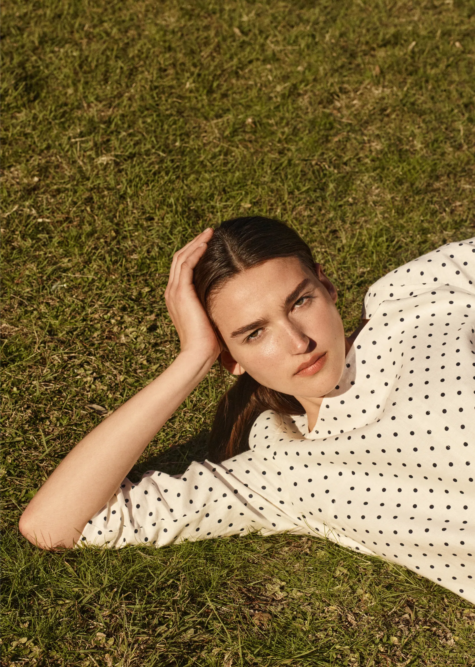 Woman lying on grass, wearing a polka dot shirt, looking at the camera.