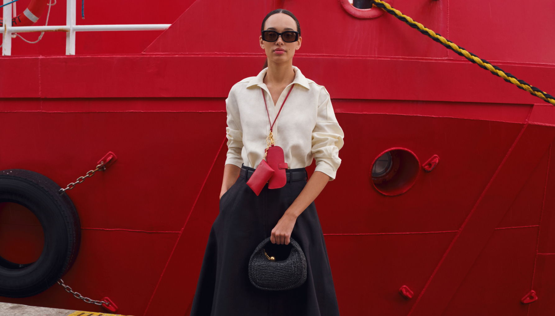 Woman in sunglasses and stylish outfit stands in front of a red boat.