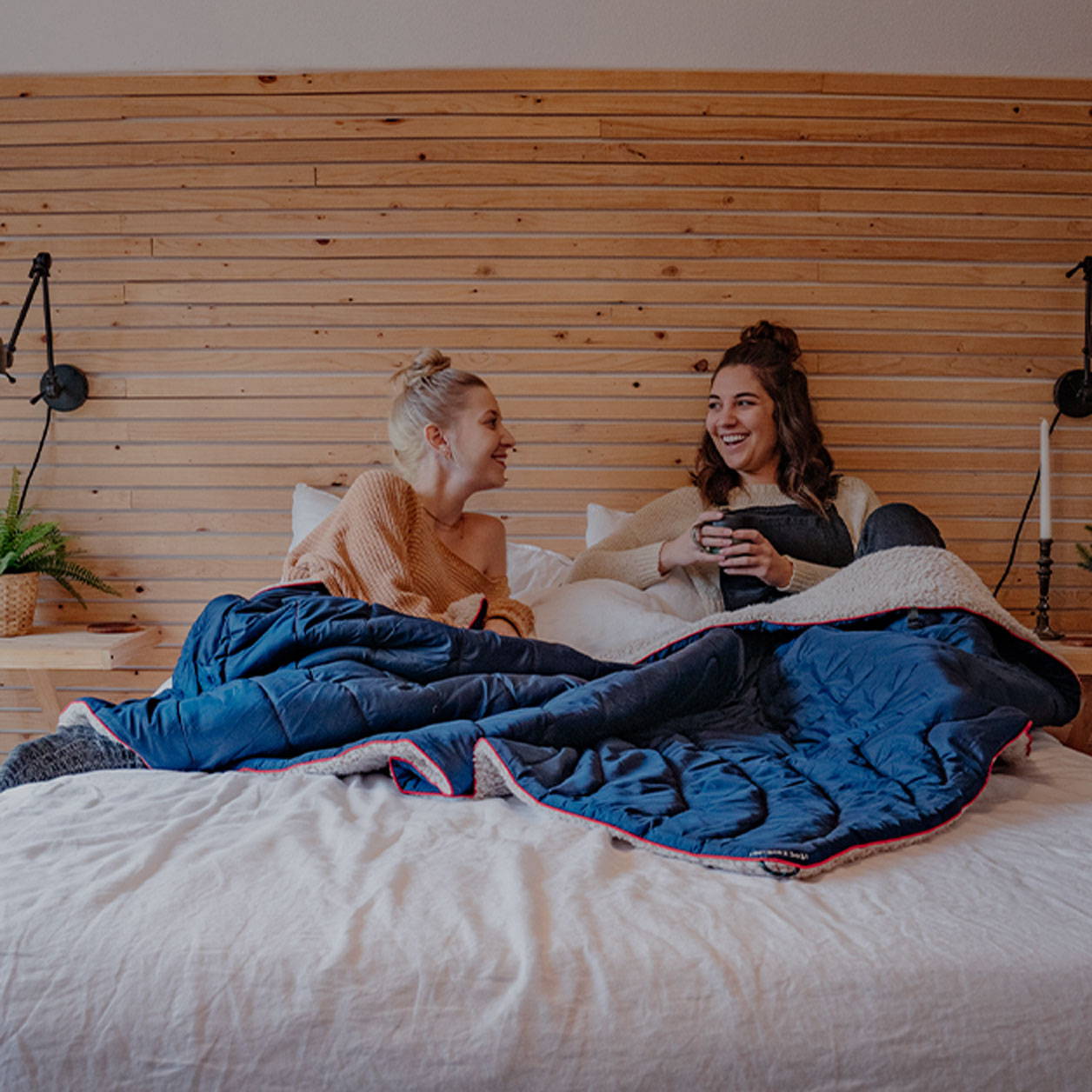 Two women laying in bed talking and drinking coffee with a sherpa fleece Rumpl blanket