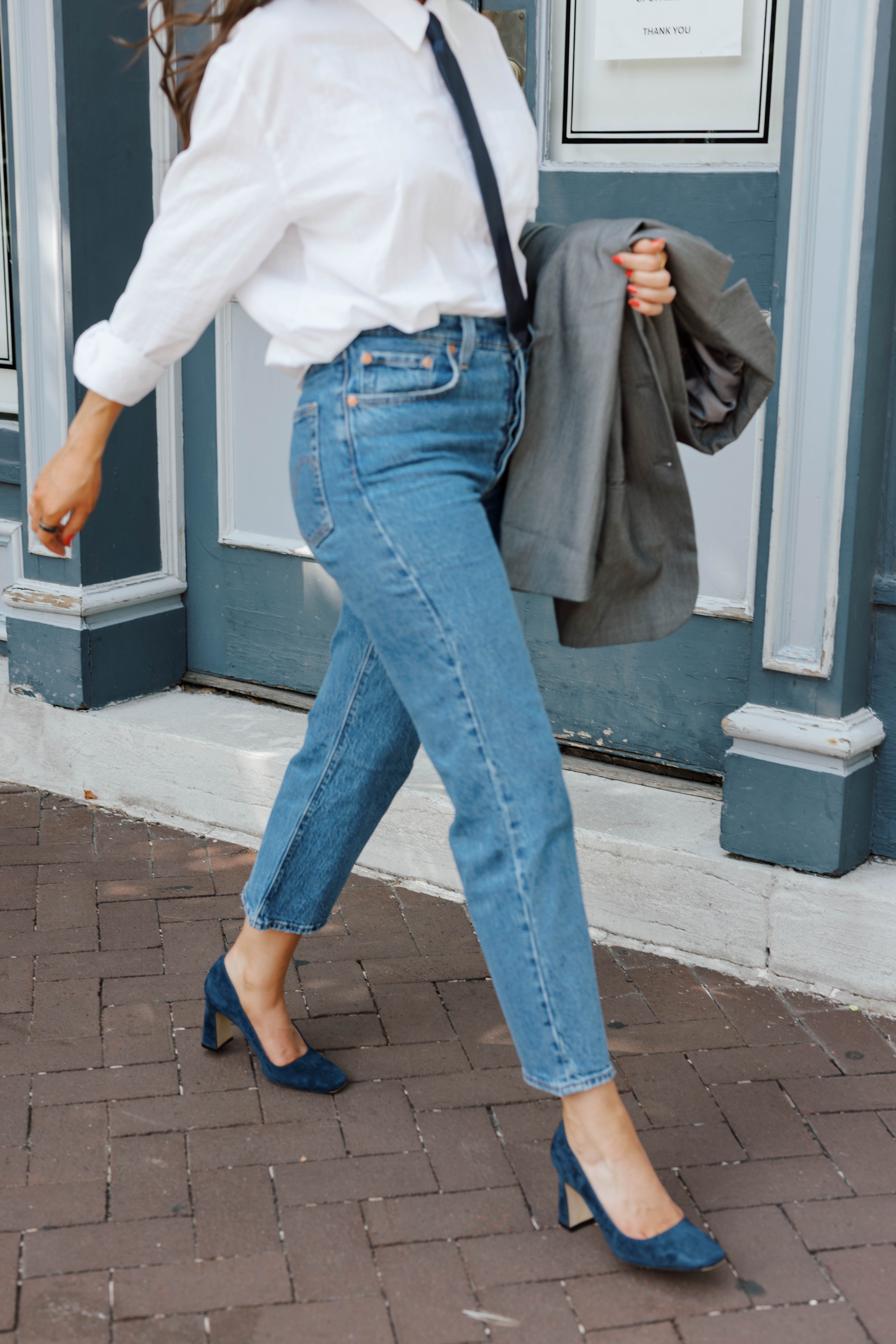 Person walking in high-waisted jeans, white shirt, and heels, holding a blazer.