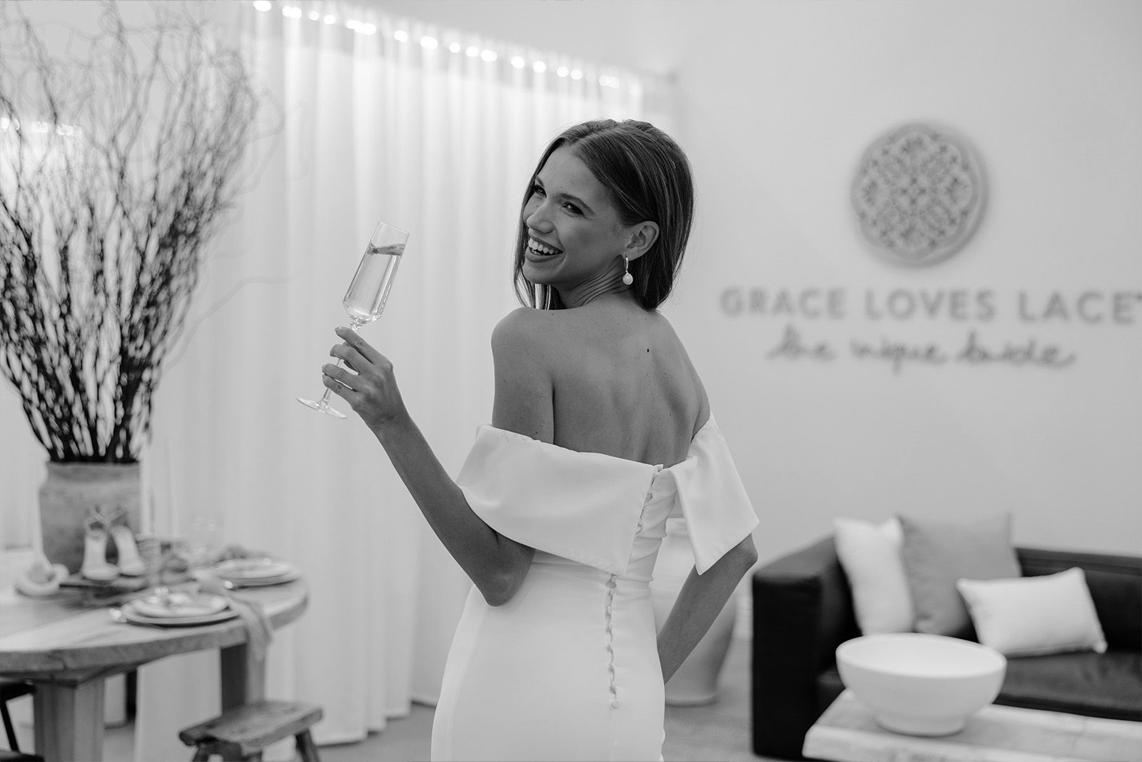 Woman in white dress holding a drink, smiling in a cozy interior setting.