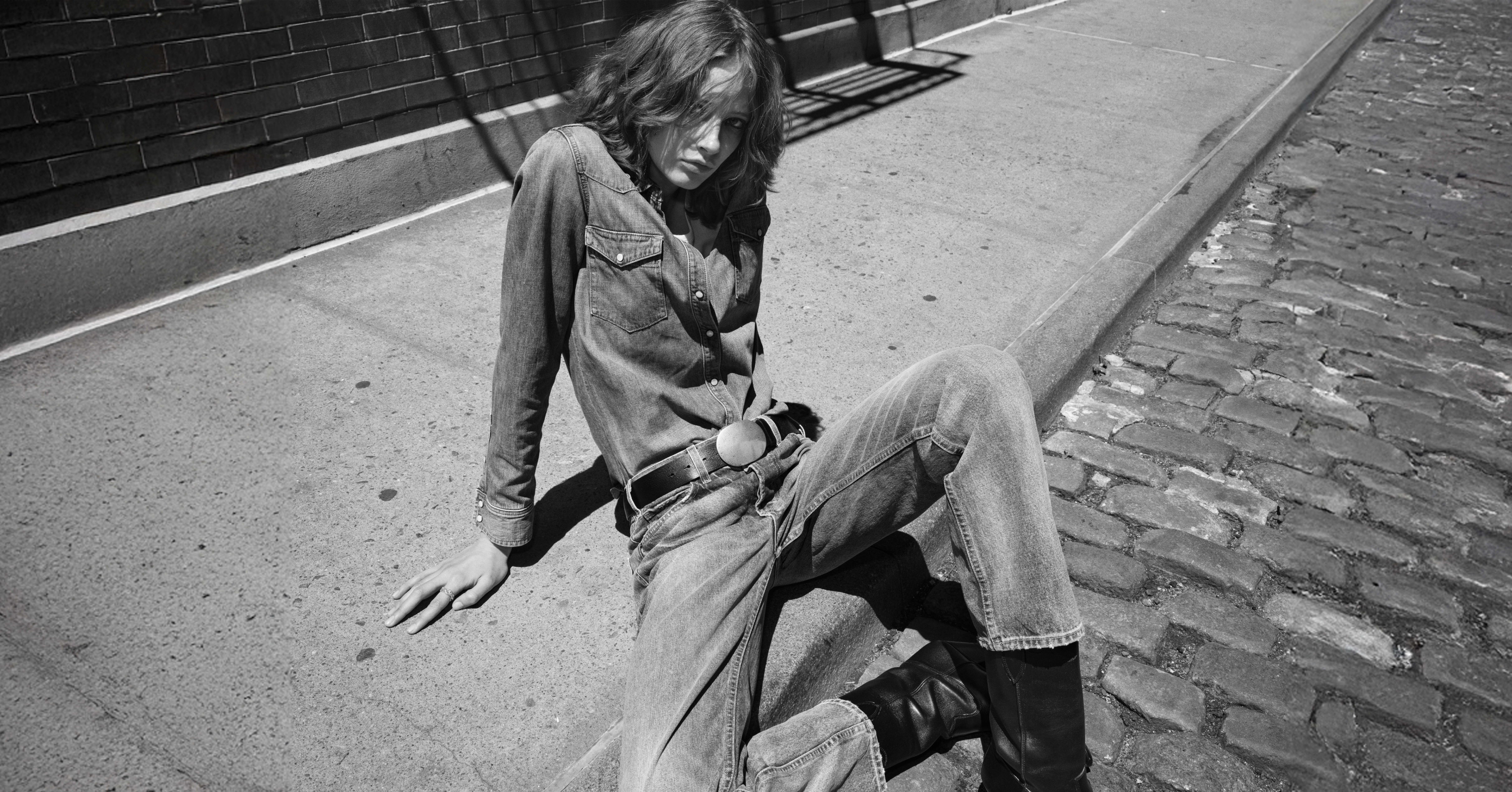 Person sitting on pavement, wearing denim outfit and boots, in black and white.