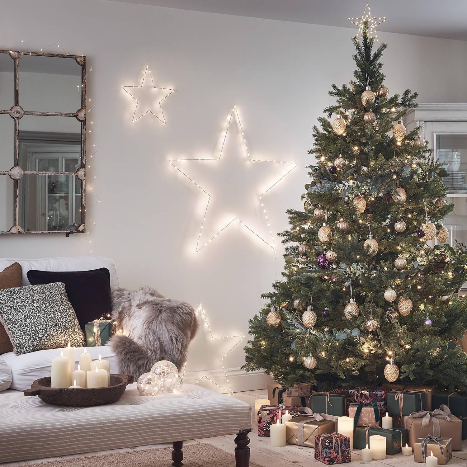 A living room decorated with a Christmas tree, star lights, LED candles, presents and fairy lights.