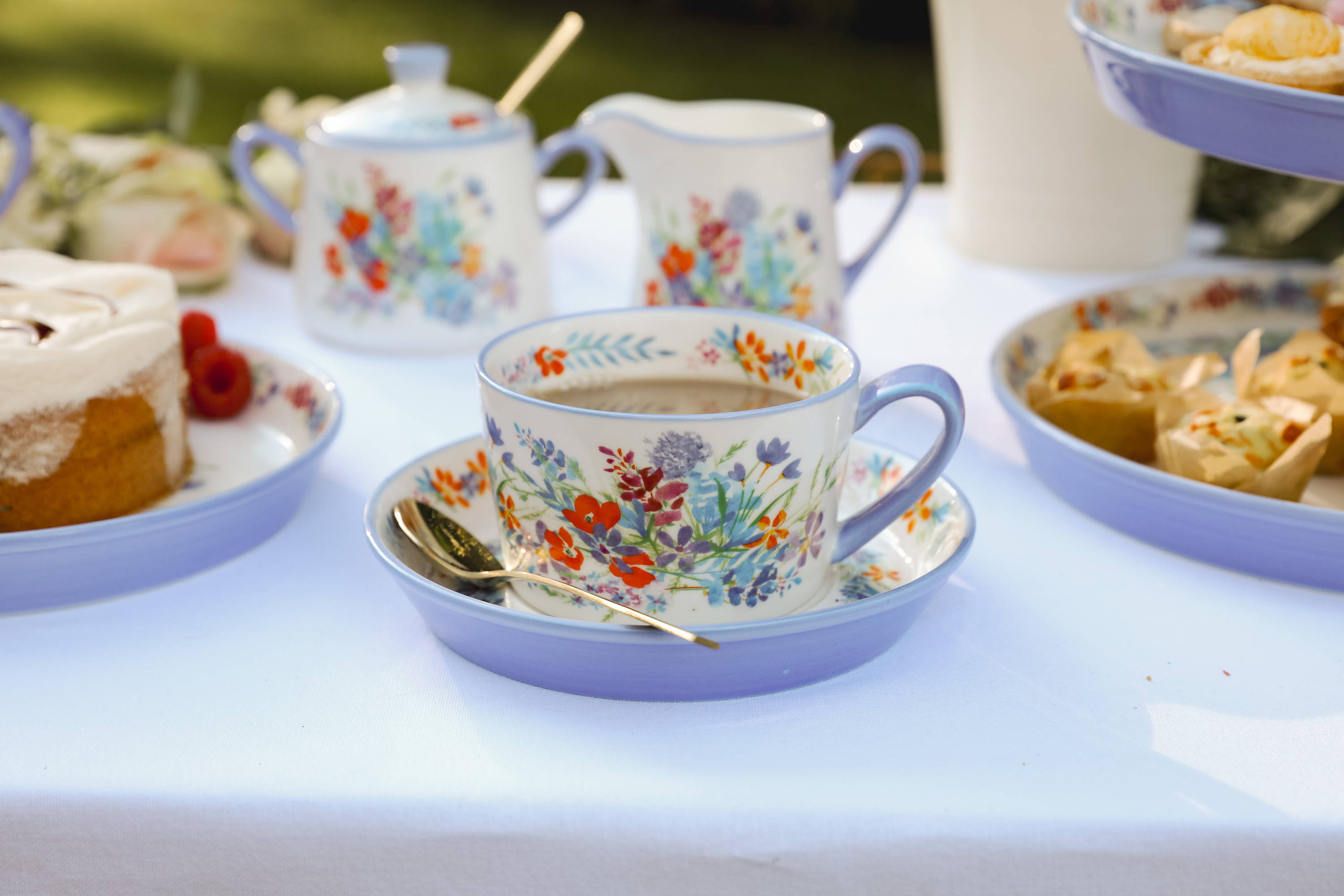 A tea cup and saucer on a table outside, surrounded by cakes and food.
