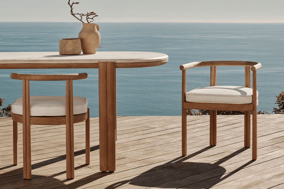 Teak outdoor dining chairs and rounded table overlooking the ocean from a coastal deck