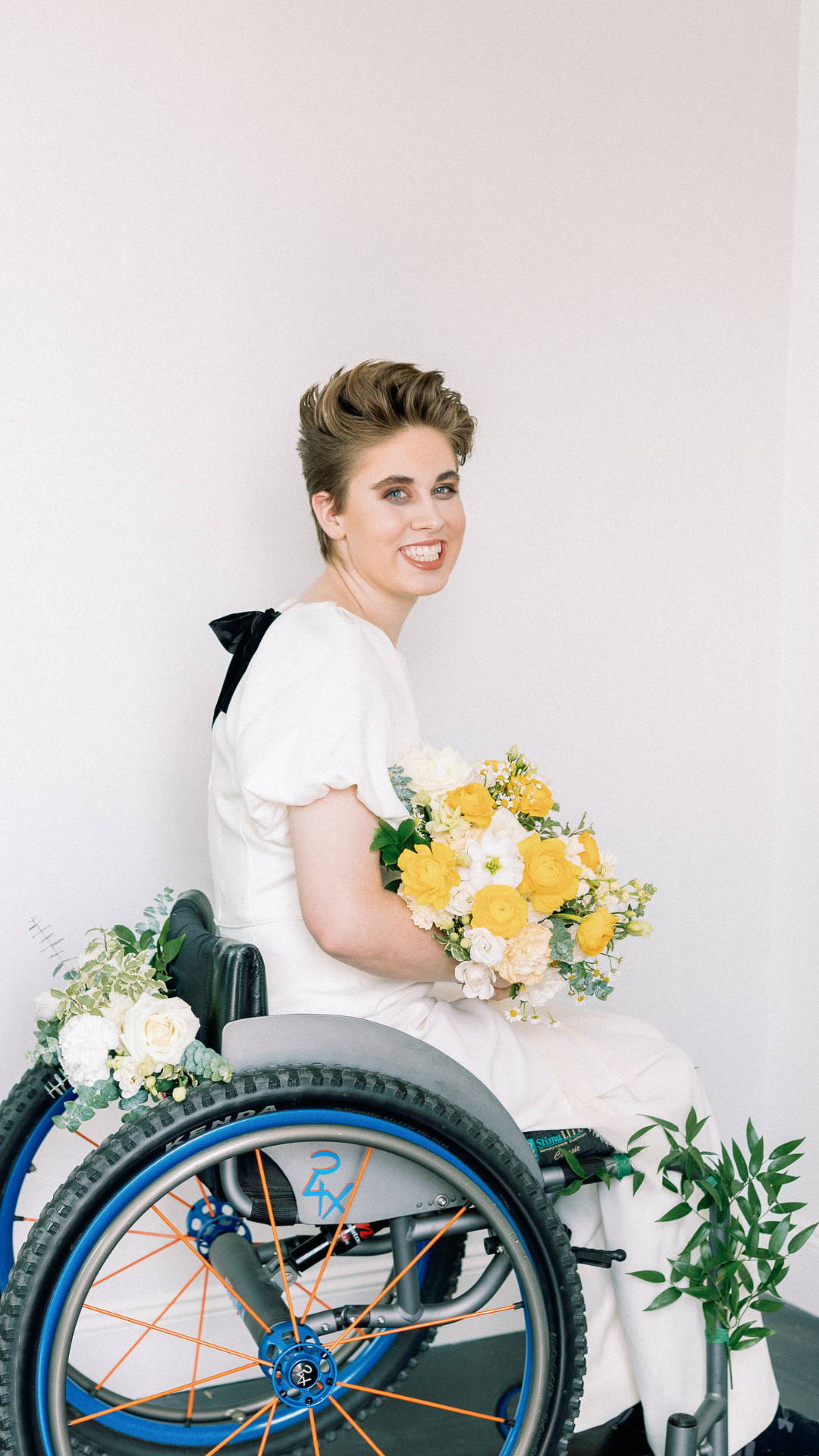 A bride in a wheelchair smiling and holding yellow flowers. 